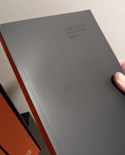 A thin black book with an orange spine. In black type at the top right of the book, only visible because of the angle the light is reflecting off it, is the text "Marcin Wichary / Shift Happens / Extras".