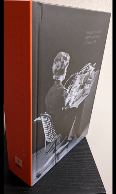 A thick book with an orange spine and a black-and-white picture of a woman with arcs of light coming from her fingers as she types on a keyboard. In black varnished type at the top right of the front cover, only visible because of the angle the light is reflecting off it, is the text "Marcin Wichary / Shift Happens / Volume One". Similarly, on the orange spine are barely-visible glyphs that reflect the light.