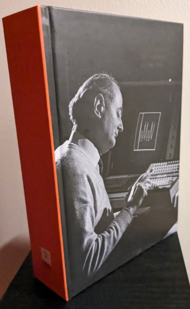 A thick book with an orange spine and a black-and-white picture of a man typing on a keyboard with a screen displaying a graph to his left. The volume is at a slightly different angle from the previous image, showing how much the light angle affects the varnish. Barely visible black type at the top right of the front cover reads "Marcin Wichary / Shift Happens / Volume Two". Similarly, the glyphs on the orange spine are almost invisible.