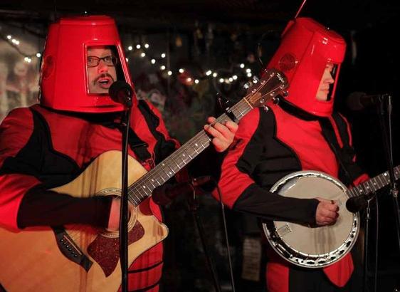 A guitar player and a banjo player standing side by side on a stage. Both are dressed in "space-y" red and black uniforms and are wearing red buckets on their heads (with holes cut out for their faces).