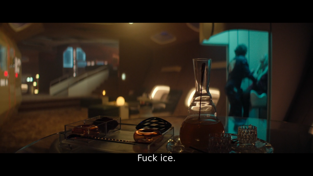 A large room inside a spaceship, decorated in earth tones with a decanter of alcohol on a table in the foreground. Two humanoid characters are visible in an alcove lit in blue-green at the right of the middle background. The captioned dialogue reads "Fuck ice."